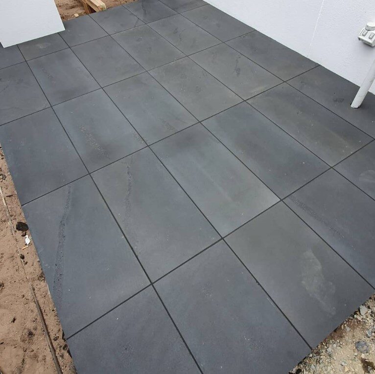 Modern grey tiles laid on outdoor patio, contrasting with unfinished dirt and gravel area.